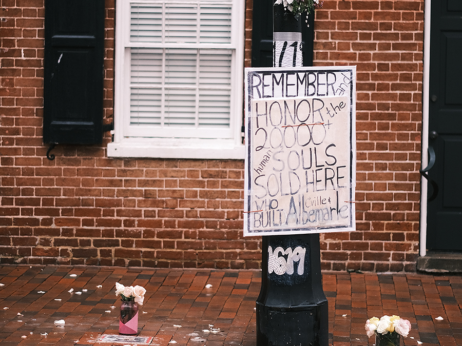 White poster that says Remember and honor the 2,000 human souls sold here who built C’ville and Albemarle.