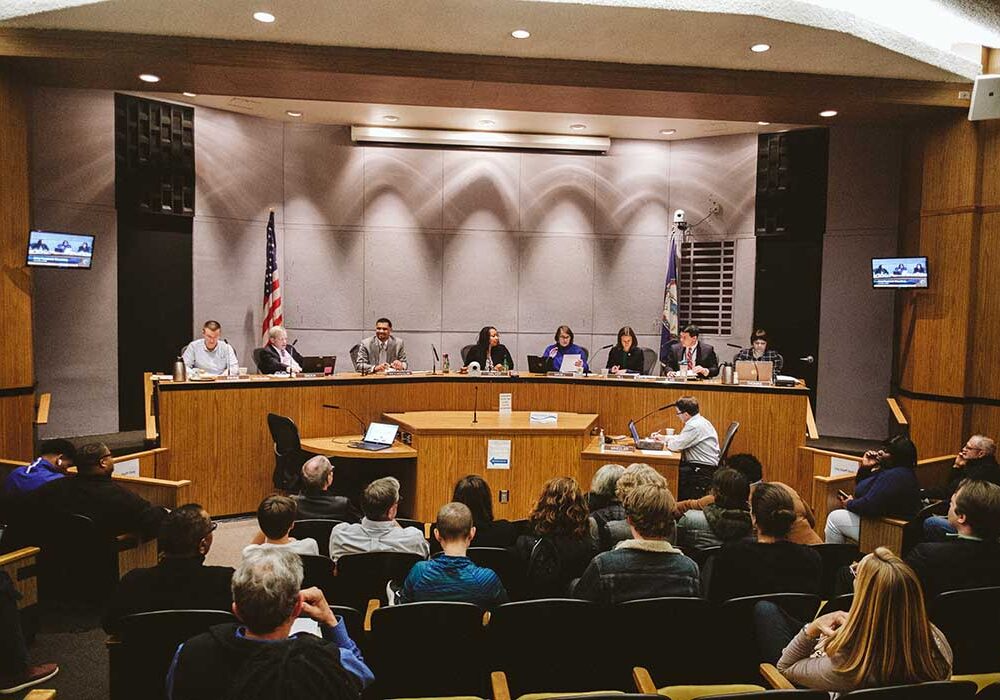 Far away photo of Charlottesville City Councilors sitting on the dais during a pre-COVID meeting inside City Hall. Around a dozen people sit in the audience, facing the councilors