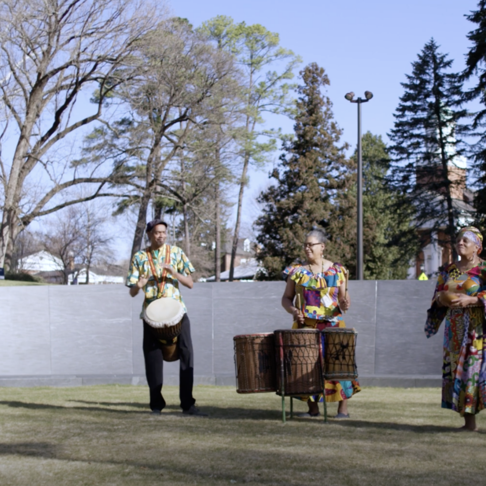 Two Black woman and one Black man play traditional drums on a green lawn in front the granite walls of the Memorial to Enslaved Laborers.
