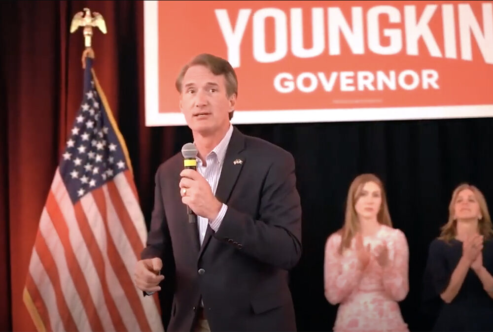 Glenn Youngkin stands on a stage with an American flag campaign sign behind him. Two women clap in the background.