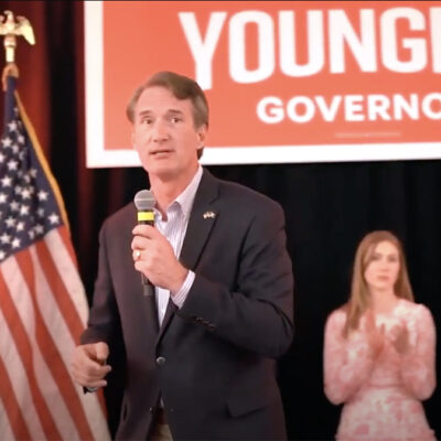 Glenn Youngkin stands on a stage with an American flag campaign sign behind him. Two women clap in the background.