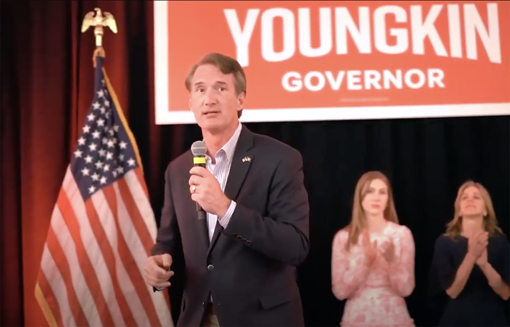 Glenn Youngkin stands on a stage with an American flag campaign sign behind him. Two women clap in the background.